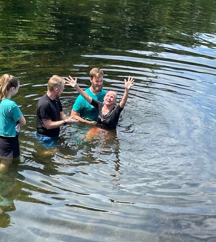 A woman raises her hands in joy during her baptism, surrounded by people supporting her in the water