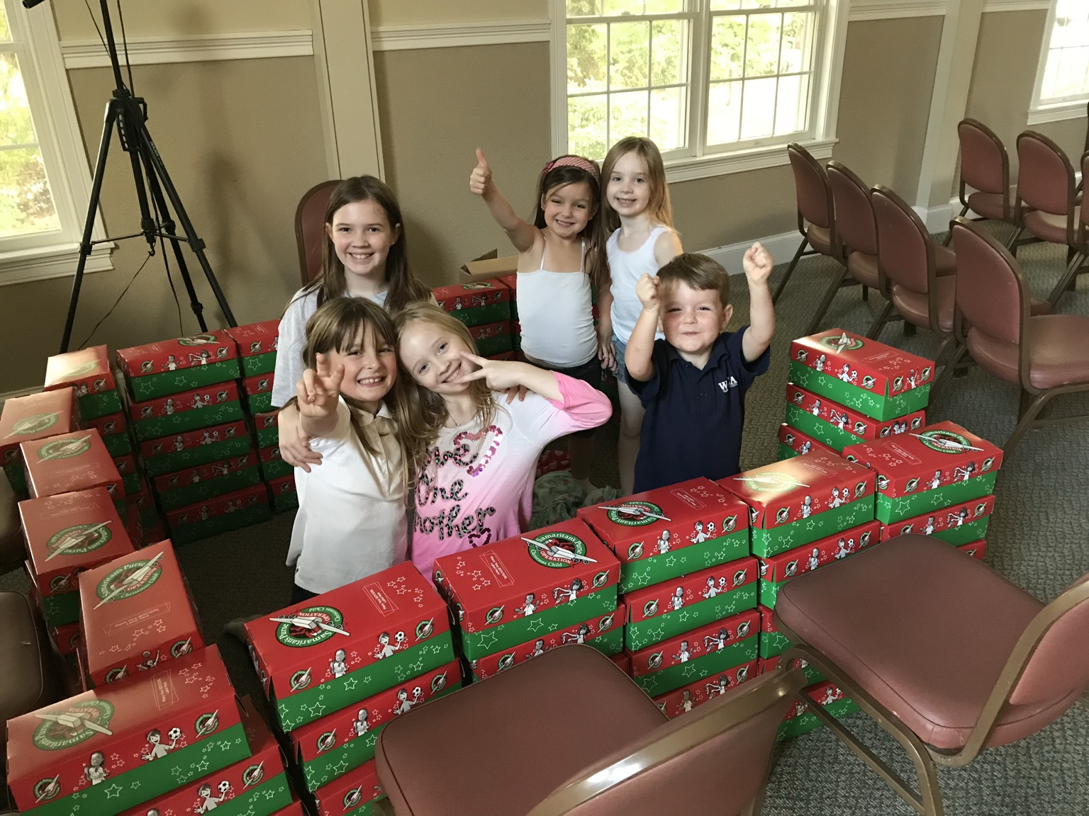Living Faith children smiling among stacks of Operation Christmas Child shoeboxes.